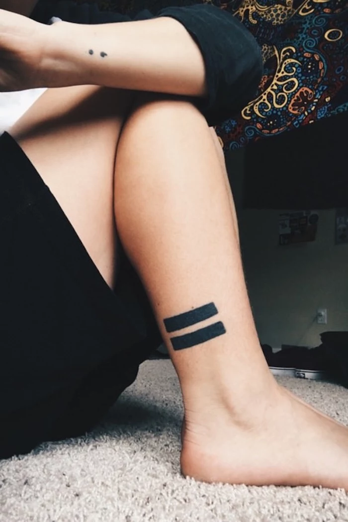 ankle with bold black tattoo, consisting of two thick stripes, and arm with semicolon tattoo on wrist, also in black, sitting person in dark clothing