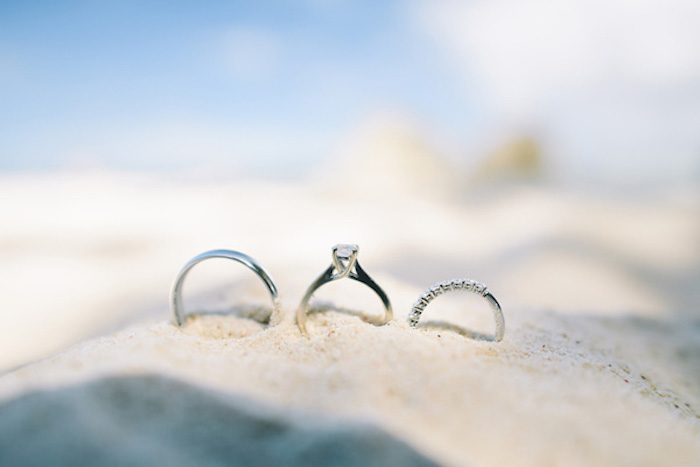 three silver rings, one thin and plain, one featuring a single diamond, and one encrusted with small stones, beach wedding ideas, placed on top of a pile of fine sand