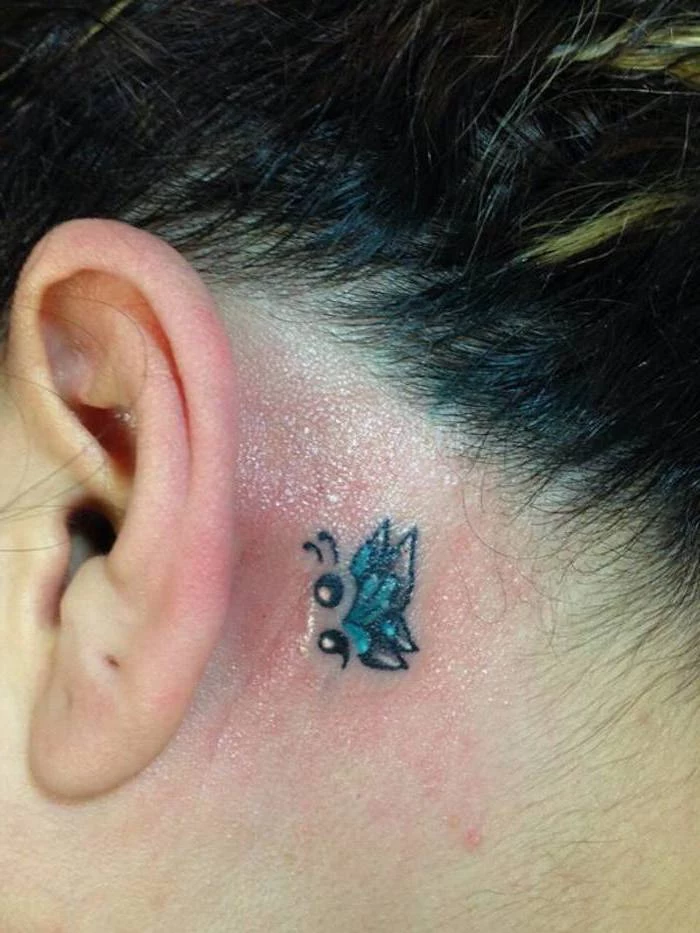 teal and black, tiny fresh butterfly tattoo, with a semicolon body, and white details, behind the ear of a woman, with dark hair and blonde highlights