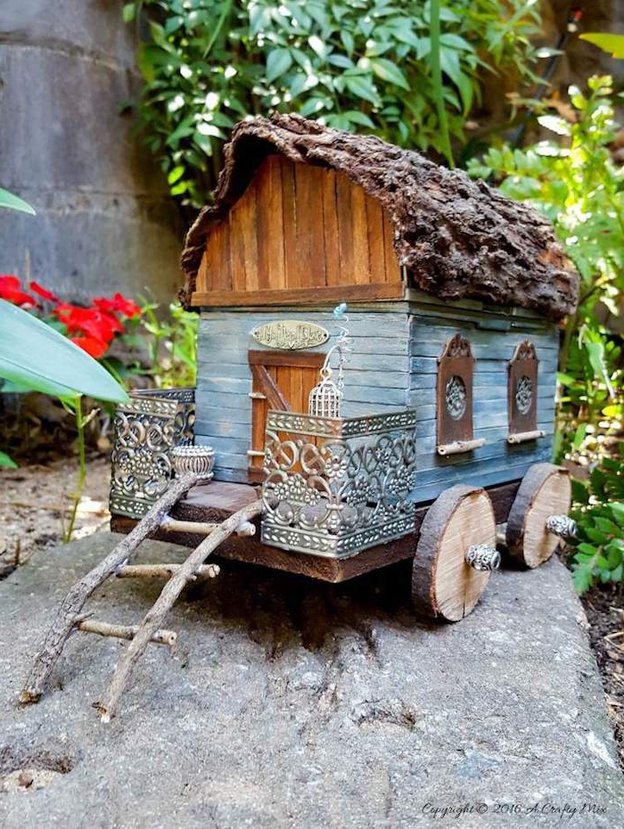 wagon or traveller's diy fairy house, made from tiny, painted wooden planks, bark and sticks, little metal ornaments, garden ion the background