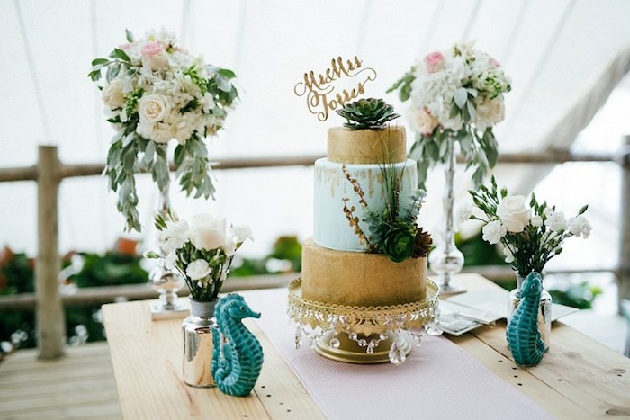 gold and pale blue wedding cake, decorated with succulents, on a wooden table, with two seahorse statuettes, and four vases, containing bouquets of different sizes