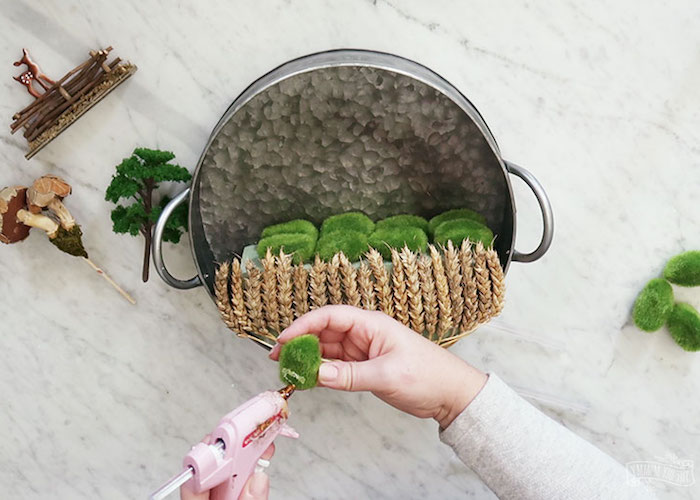 applying glue on a piece of moss, using a pink glue gun, placing the moss on top of a sponge, decorated with dried wheat stalks, fairy garden inside a metal cooking pot