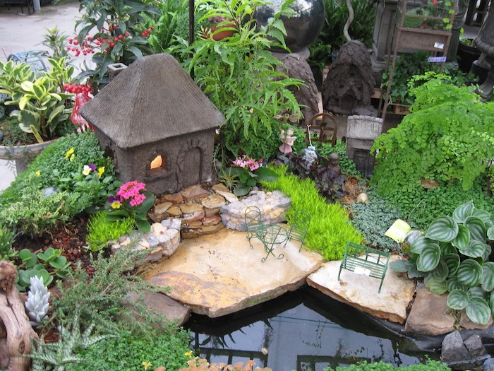 artificial pool surrounded by stones of various sizes, in a garden with many different kinds of green plants, decorated with miniature fairy house made of stone, a tiny bench, and a small table with matching chairs
