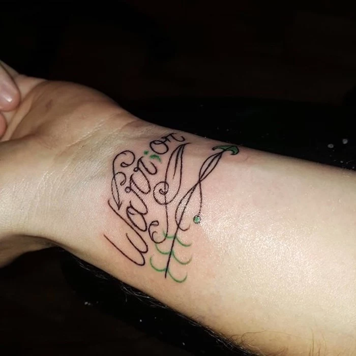 the word warrior, written in a fancy black font, with the letter i replaced by a green semicolon, tattooed on a person's wrist, semicolon tattoo meaning, arrow decorations in black, and light green