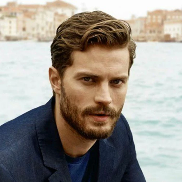 jamie dornan with wavy, short hair and bangs combed over to one side, beard and mustache, dark denim blazer and blue t-shirt