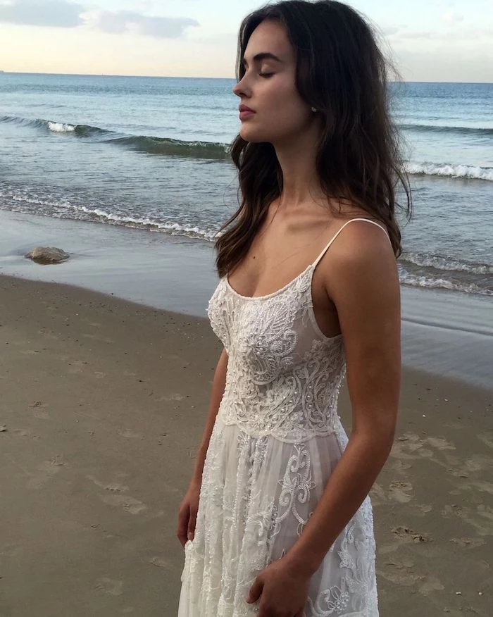 strappy white maxi dress, with embroidery and sequins, on a young tanned brunette woman, with closed eyes and wavy hair, standing on the seashore
