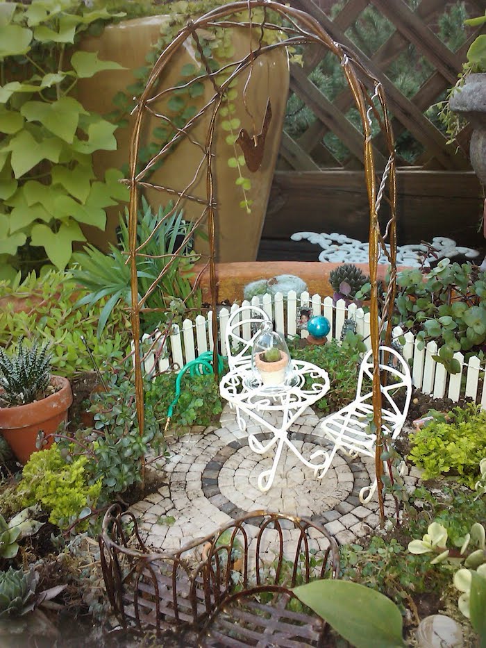 wire arch decoration, inside a miniature garden, with tiny white wrought iron table and matching chairs, tiled floor and picket fence, fairy garden pictures, various small green plants