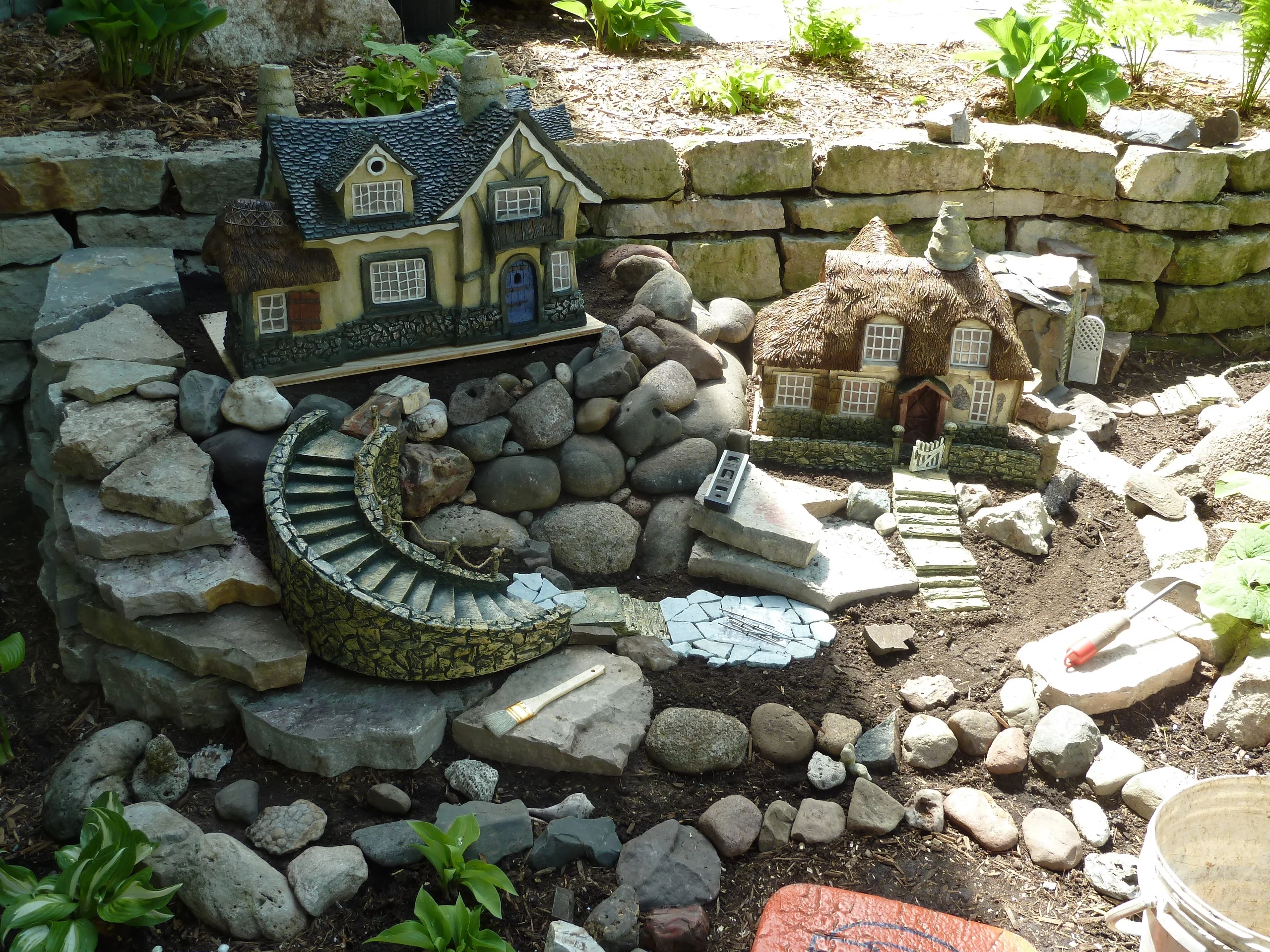 fairy garden pictures, garden with lots of gray stones of different sizes, decorated with two miniature houses, several green plants and tools