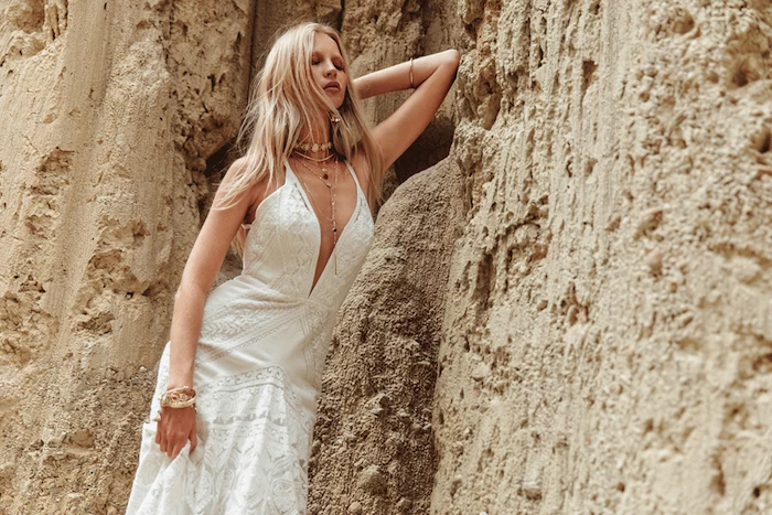 platinum blonde woman, wearing a white lace boho wedding dress, with several necklaces and bracelets, beach wedding attire, leaning on a pale beige rock