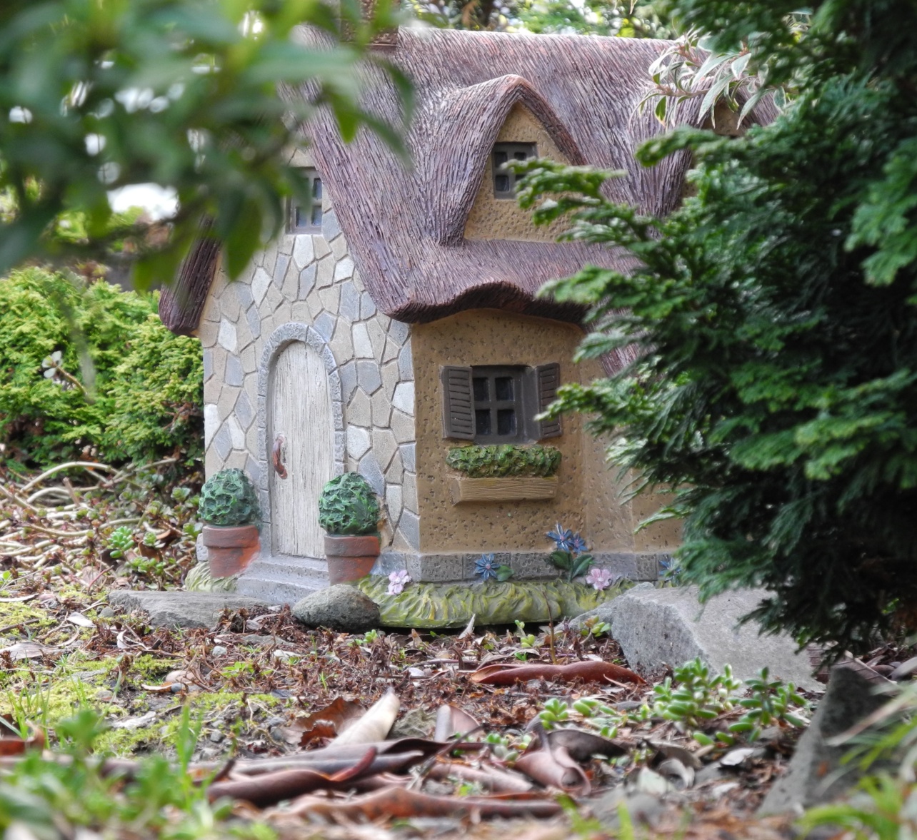 house-shaped garden decoration, made from ceramic or moulding clay, placed on a forest floor, diy fairy house, near green shrubs and branches