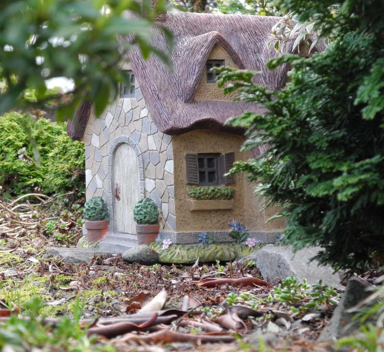 house-shaped garden decoration, made from ceramic or moulding clay, placed on a forest floor, diy fairy house, near green shrubs and branches
