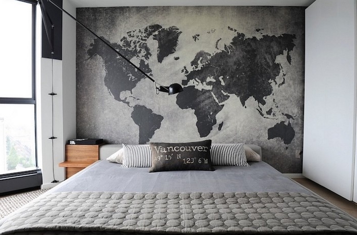 large map of the world, in dark and light gray, on a wall near a simple double bed, with several cushions, bedroom design, large window on the left
