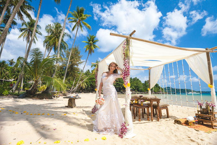 happy bride in long, cream and white embroidered wedding gown, leaning on a beach tent, decorated with flowers, beach wedding venues, palm trees and sea, wooden furniture and sand
