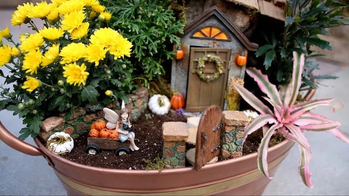 autumn-themed fairy garden, inside a metal bucket, with yellow flowers, and a pink and green air plant, decorated with small house, and a figurine of a fairy, sitting on a miniature pumpkin cart