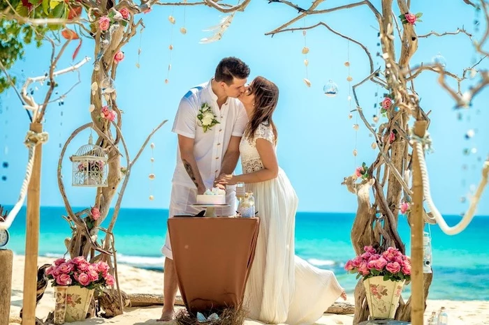 lots of pink roses, and a wedding arch formed by intertwining trees, on a white sandy shore, near an azure blue sea, beach wedding venues, kissing bride and groom
