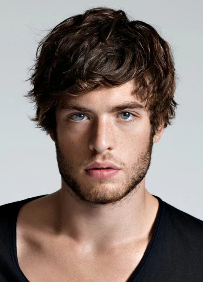 boyish layered haircut, with wavy bangs falling on the forehead, guys with curly hair, blue-eyed man with short beard, wearing a black slouchy top
