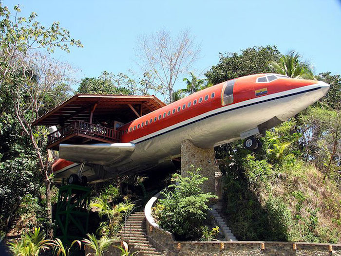 red and silver airplane, placed on stone pillars, and supported by trees, containing an adult treehouse, with wooden terrace and roof