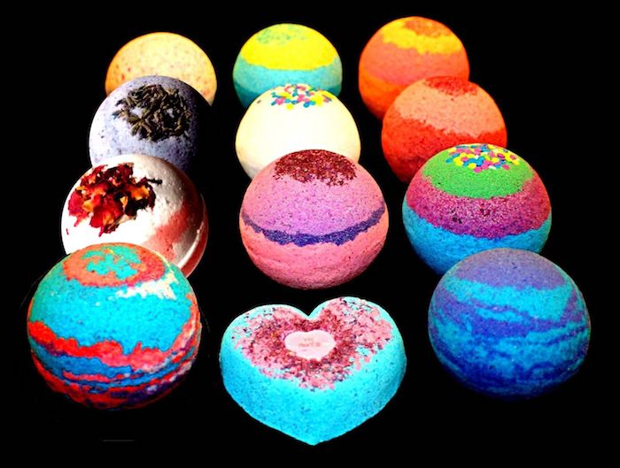 twelve different bath bombs, various colors and toppings, some are striped, one is in the shape of a heart, on a black background