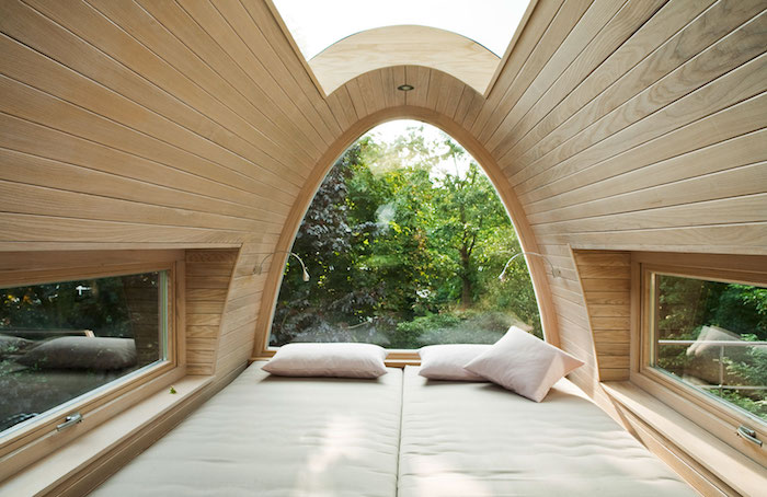 three pillows on a double bed, inside a capsule-like construction, with wood paneling, and three windows, treehouse designs