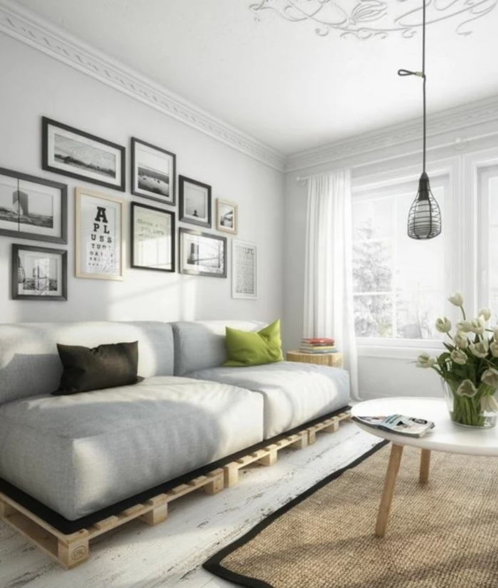 nordic style room, with white floor, walls and ceiling, containing a modern coffee table, a diy pallet sofa, a beige rug, and a hanging lamp
