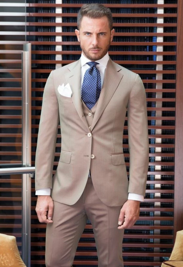 tight-fitting three piece suit in beige, worn with a white shirt, dark blue patterned tie, and a white pocket handkerchief, how to dress for a wedding male, serious looking man