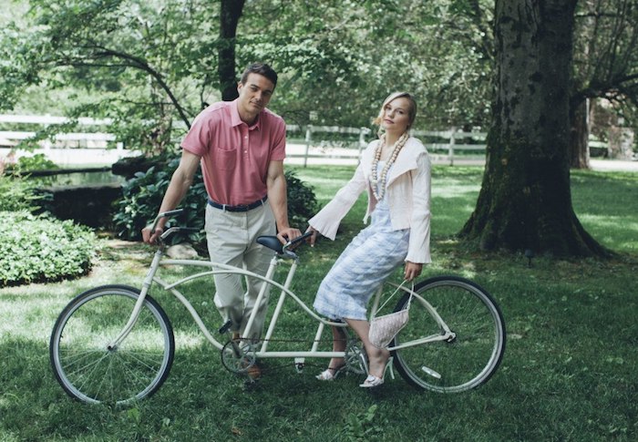 couple with a bicycle for two, man with pink polo shirt, and off-white trousers, leaning on the handlebar, dressy casual men, woman in pale blue and white jumpsuit, sitting at the back, garden chic wedding dress code
