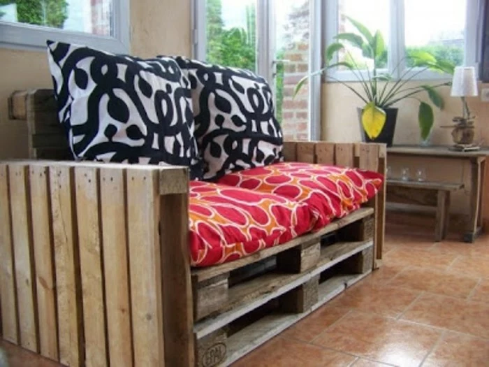 cushions in black and white, and orange and red, decorating a diy sofa, made from wooden pallets, inside a bright room, with a small table and potted plant