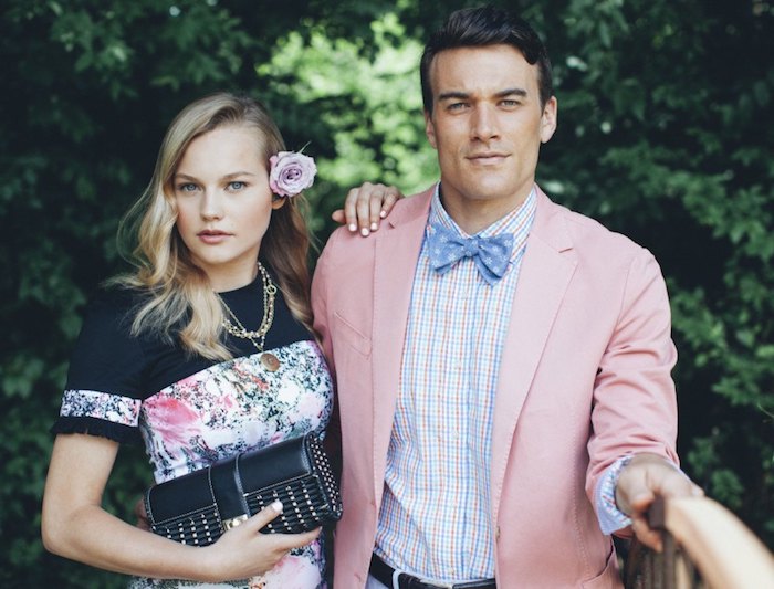 dressy casual men, blonde woman in black top, with floral details, leaning on man in pale pink blazer, worn over white, pale pink and pale blue checkered shirt, wit blue patterned bowtie