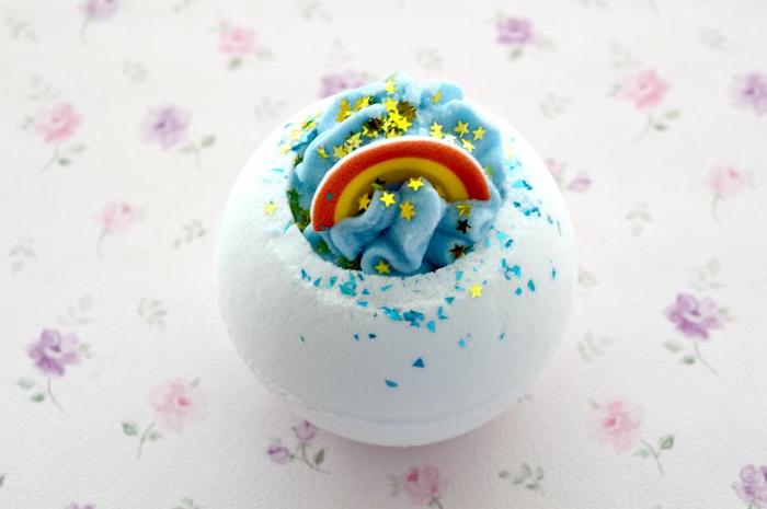 rainbow bath bomb, white and blue, decorated with red and yellow bow, tiny golden star-shaped confetti, and blue sprinkles, how to use a bath bomb, placed on floral cloth