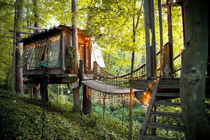 fairy lights illuminating a boho-style tree house, treehouse designs, built around several trees, and connected to a nearby structure via a bridge 