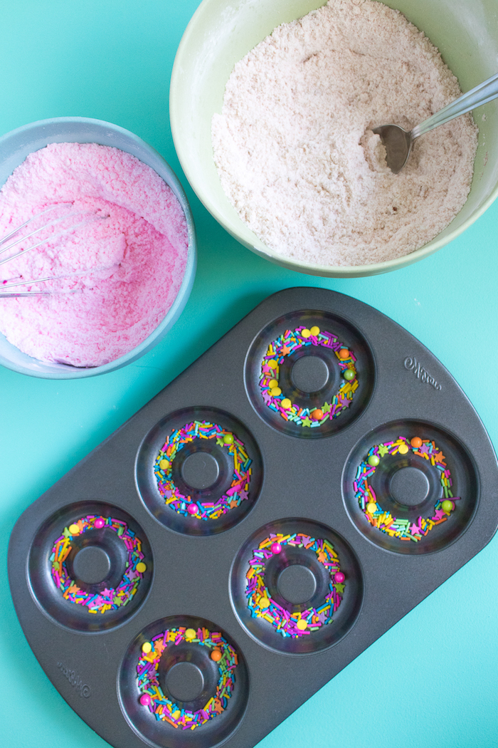 pale pink powder, in baby blue bowl, near bigger light green bowl, containing fine white powder, and a metal spoon, donut shaped bath bombs recipe