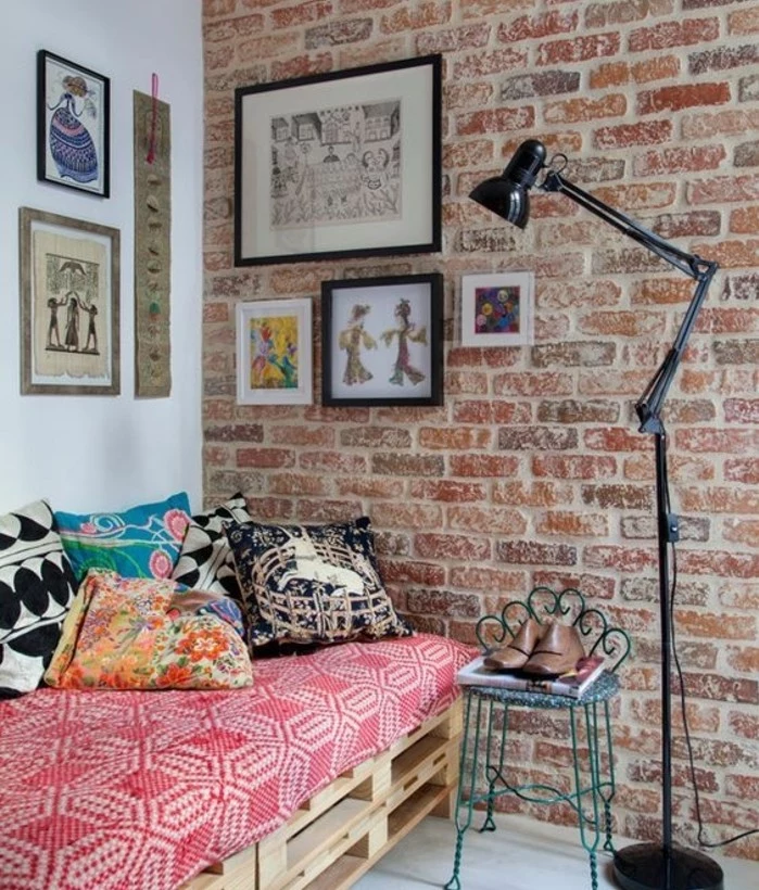several artworks decorating a room with one white wall, and one brick wall, containing a pallet settee, with lots of cushions, how to make pallet furniture, tall lamp and a vintage chair