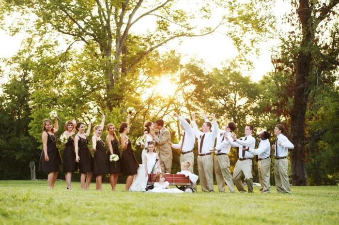 wedding photo on a green lawn with trees, bride and groom kissing, flower girls and bridesmaids, six men in coordinated outfits, how to dress for a wedding male, white shirts and beige trousers