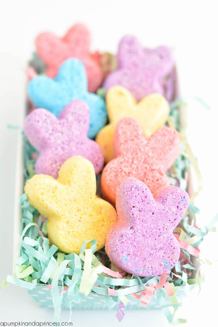 rabbit shaped bath bombs, in different pastel colors, placed in a plastic box, filled with multicolored easter grass, cartoon bunny heads