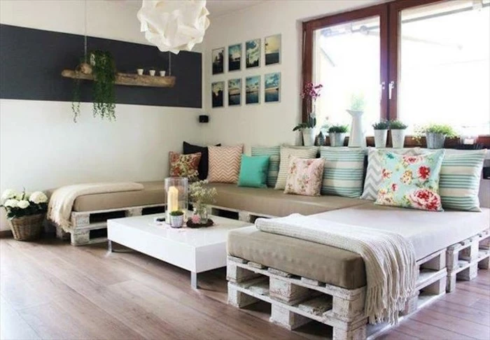pallet couch shaped like a c, covered with beige foam mattresses, and decorated with lots of cushions, in different sizes and colors