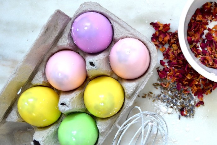six plastic easter eggs, in different pastel colors, filled with bath bomb recipe ingredients, and placed in a egg carton, near dried flower petals, and different utensils