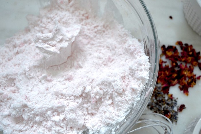 very pale pink chalky powder, inside a clear glass bowl, placed on a white surface, bath bomb recipe, dried lavender and rose petals