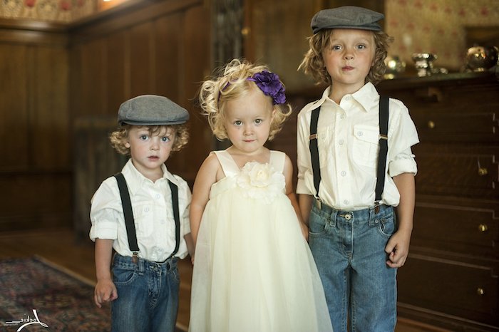 kids dressed in semi formal clothing, garden party attire, two boys wearing jeans, with white shirts and suspenders, and grey vintage berets, and a little girl in a cream dress, with a purple flower ornament in her hair 