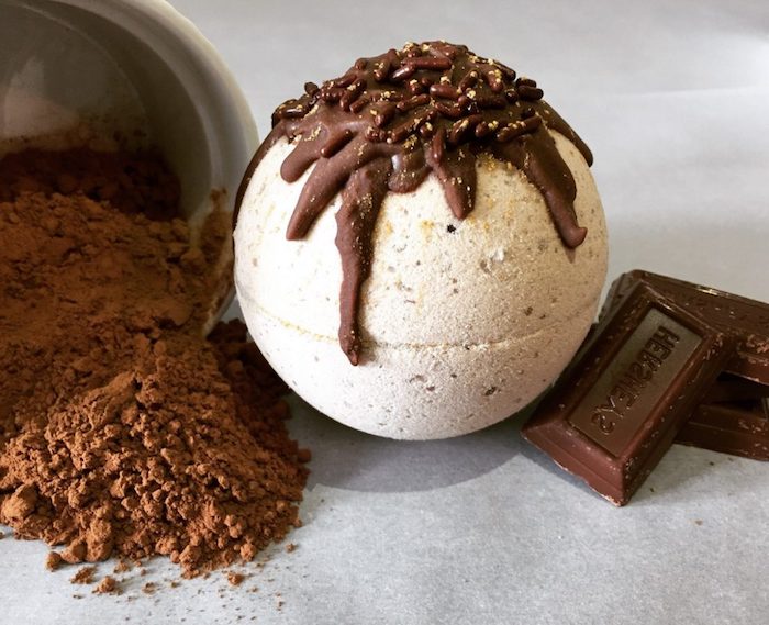 beige spheric bath bomb, decorated with chocolate-like topping, what is a bath bomb, several pieces of chocolate, and a dish with brown powder nearby