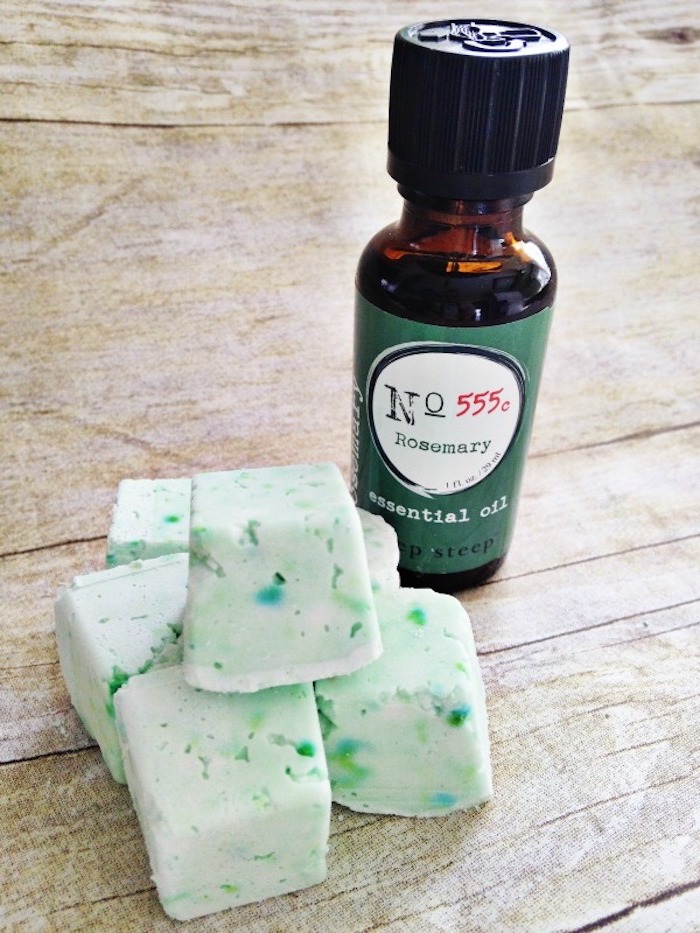 rosemary bath melts, in cubic shape, pale spotty green and white, on beige surface, bottle of essential oil nearby