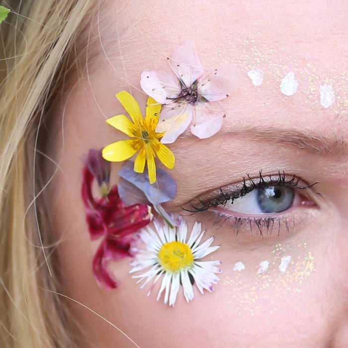 several different pressed flowers, stuck near the eye of a blonde woman, wearing black mascara, makeup looks, several spots of white paint