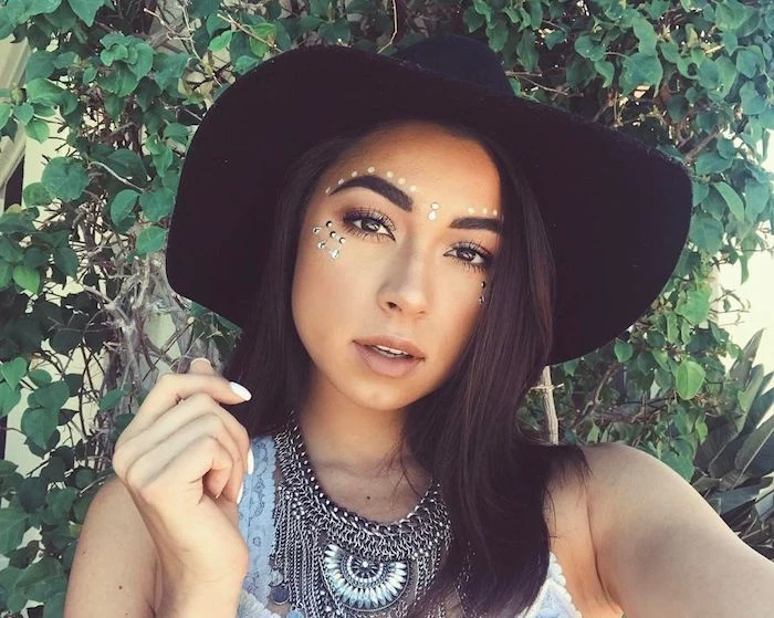 hat in black, worn by brunette woman with dark eyes, discrete and natural make up, decorated with white face paint, and silver pearl stickers, cute makeup looks, boho clothing and jewelry