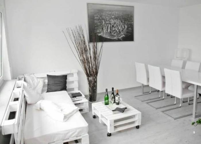 minimalistic interior in white and pale grey, modern dining table with six chairs, pallet sofa and a matching coffee table, large grayscale photo on the wall, decorative dried branches in a vase