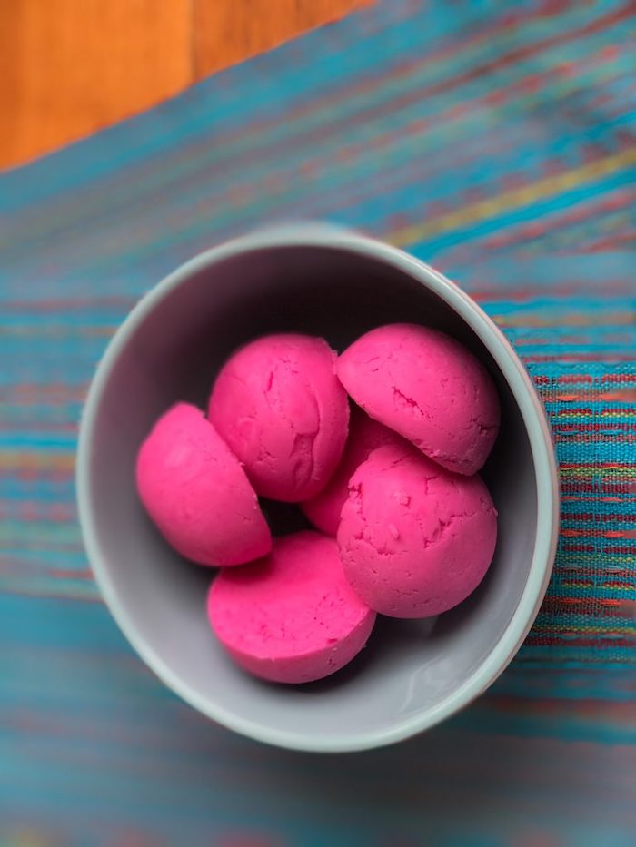 hot pink bath melts, shaped like half spheres, and placed in a pale grey ceramic bowl, on a striped multicolored cloth