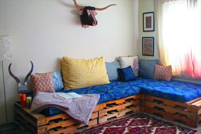 ornamental rug in red and white, near a corner pallet couch, decorated with blue patterned fabric, and several different cushions