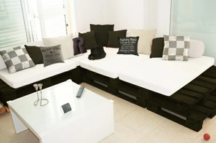 black and white corner sofa, with many cushions in grey, white and black, in different sizes, and featuring different patterns, furniture made from pallets, modern white table nearby