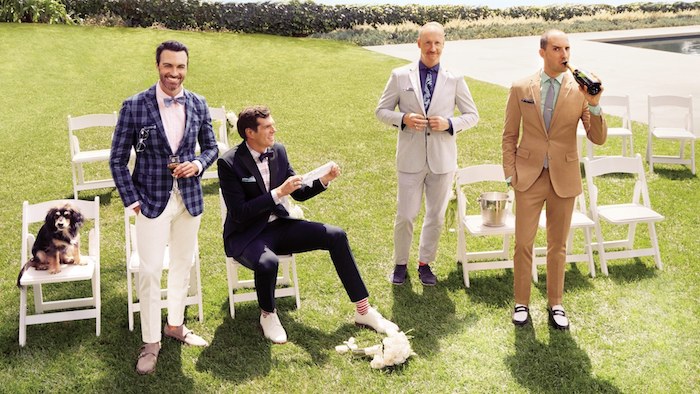 four men in semi formal clothes, and a dog, standing on a green lawn, near several white chairs, one of the men is sitting, dressy casual men, checkered blazer and white trousers, suits of different colors, garden chic wedding dress code