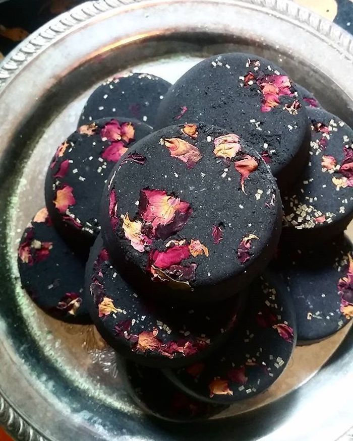 flat bath bombs in black, with dried rose petals, in yellow and different shades of pink, placed on an ornamental, round silver tray