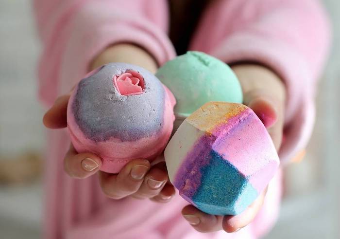 woman in pink robe, holding three bath fizzies in her hands, round and gem-shaped, pale green and multicolored