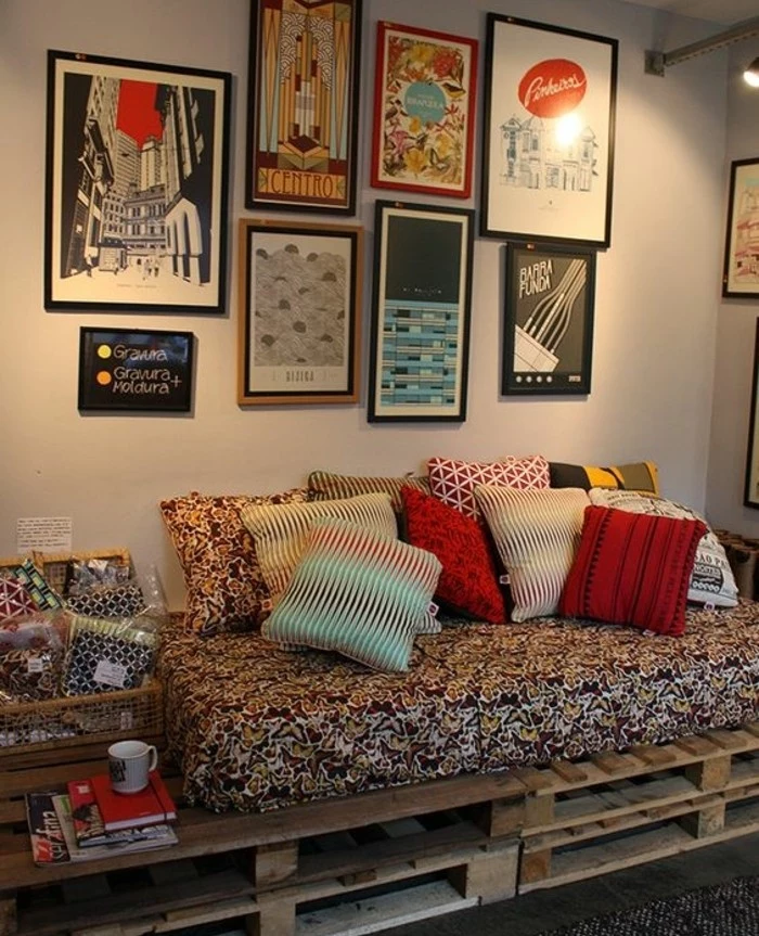 posters covering a wall, near a pallet sofa, mattress with multicolored cover, and several cushions, with different colors and patterns